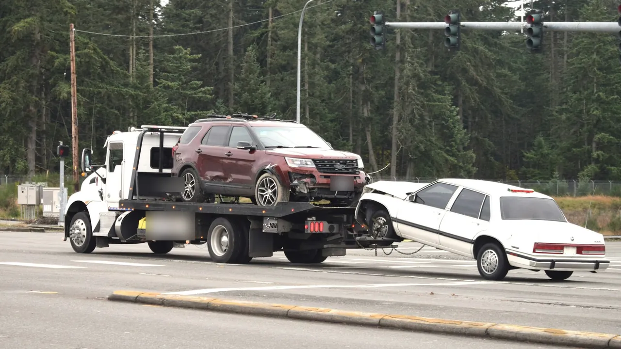 White flatbed tow truck carrying a damaged red SUV while towing a white sedan by its front wheels through an intersection