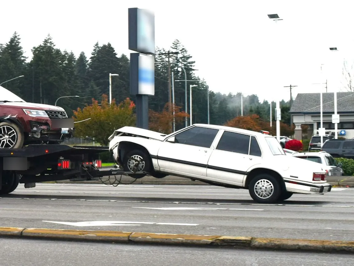 White sedan with front-end damage lifted by its front wheels behind a tow truck